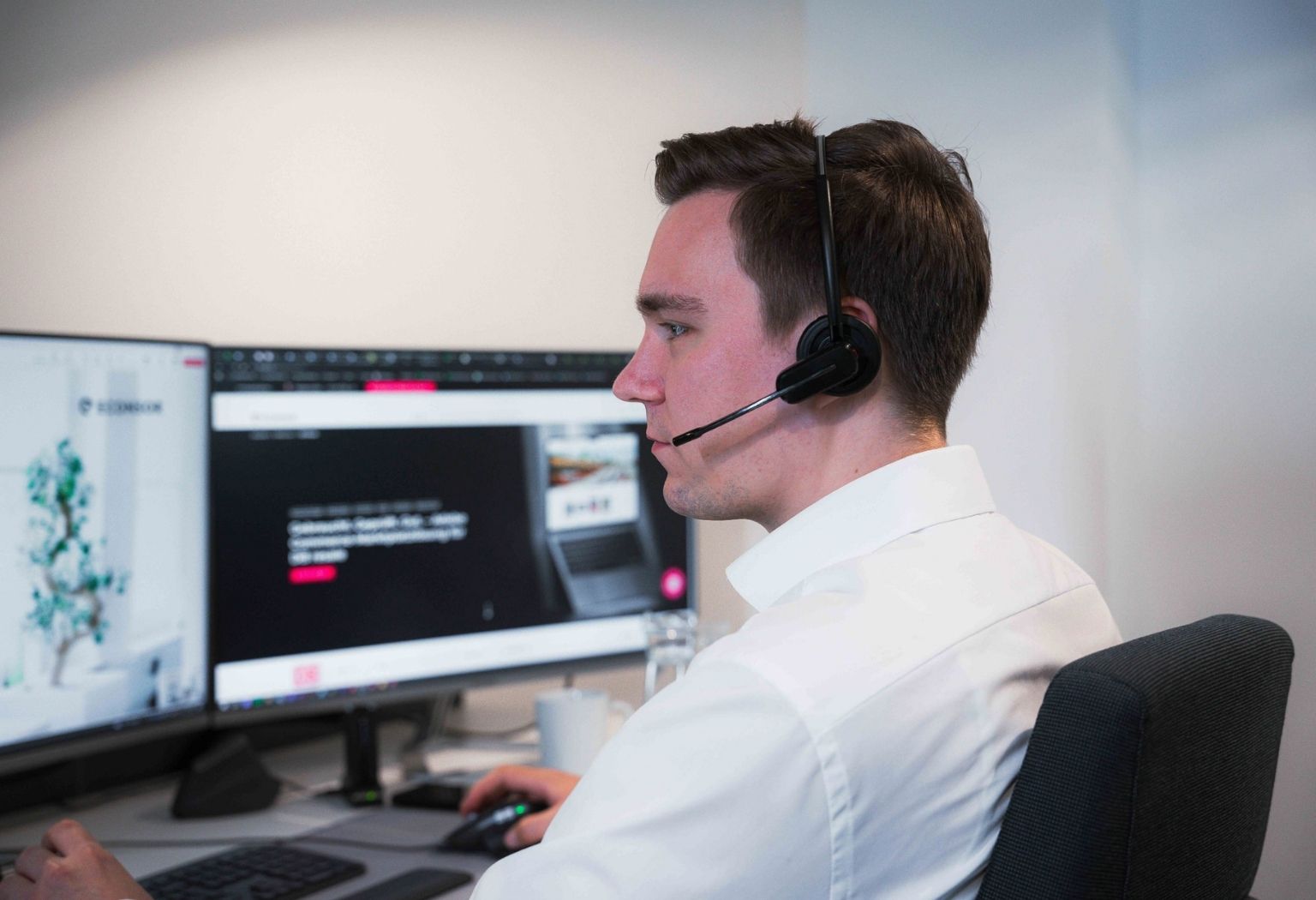 Mann mit Headset an einem Schreibtisch, umgeben von zwei Monitoren, konzentriert auf seine Arbeit.